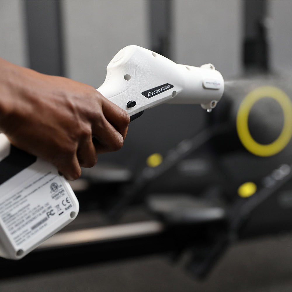 Billede af det hvide håndtag på protexus backpack elektrostatiske sprayer. Her ses håndtaget fokuseret i et fitness miljø, som er sløret. Ud af håndtaget bliver der forstøvet en væske. Håndtages holdes af en hånd.