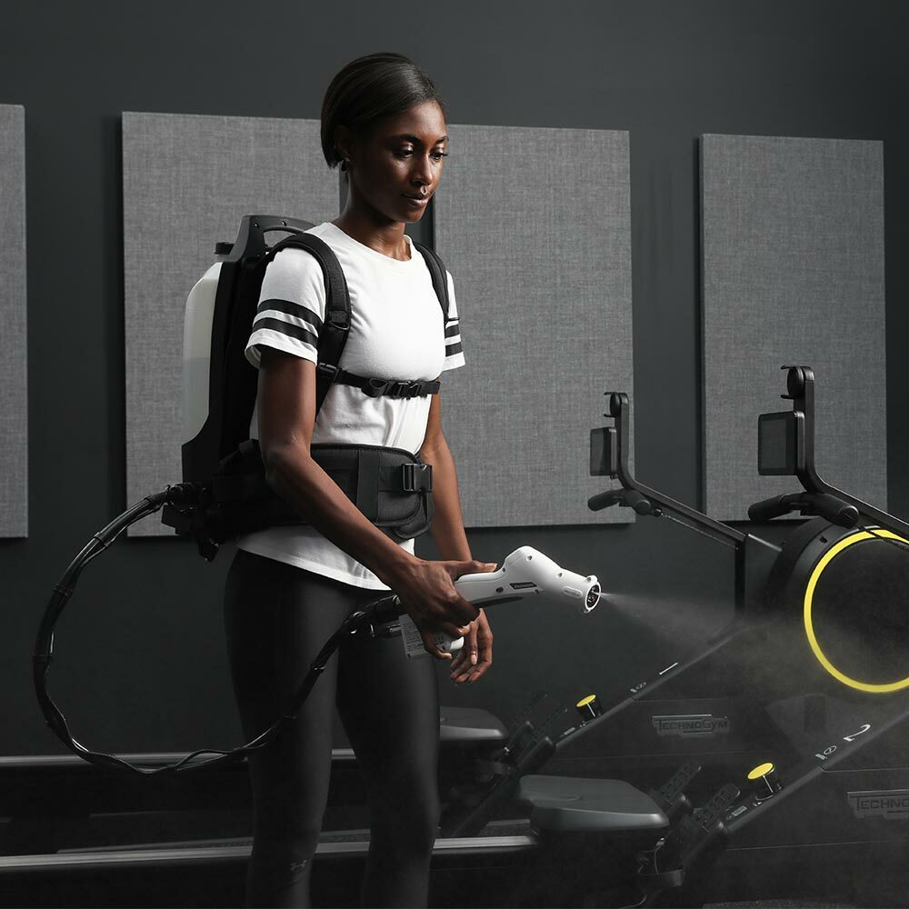 Billede af protexus backpack elektrostatiske sprayer, som anvendes til at desinficere et fitnesscenter. Sprayeren sidder på ryggen af en kvinde med hvid trøje, som holder håndtaget på sprayeren med sin højre hånd. Ud af dysen på håndtaget forstøves væske som lægger sig på overfladerne i rummet.