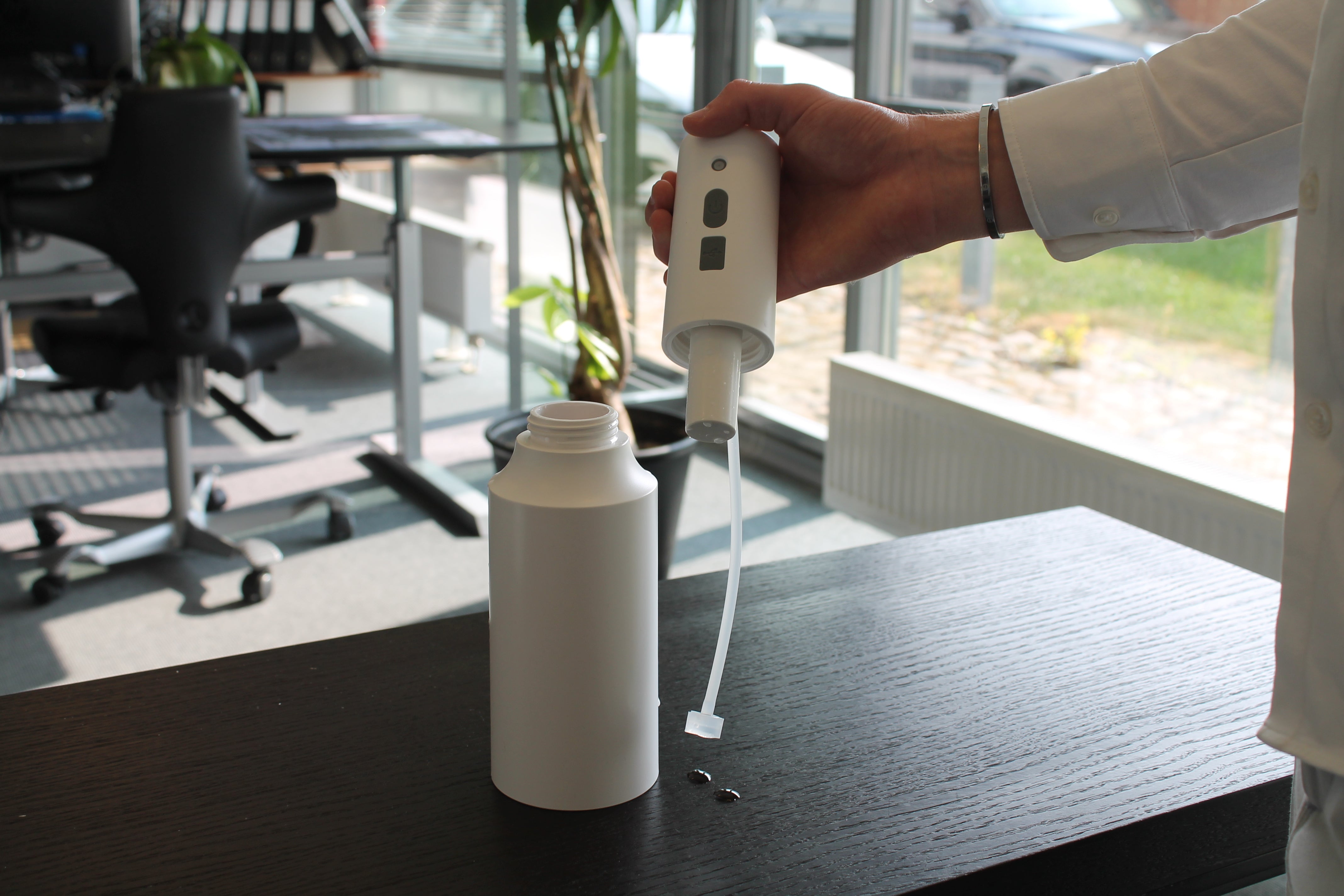 Produktbillede af safesprayer elektriske forstøver tanken og "hovedet" er taget fra hinanden. Man kan her se hvor desinfektionsmiddel skal påfyldes. Den står på et sort bord og i baggrunden er kontormiljø.