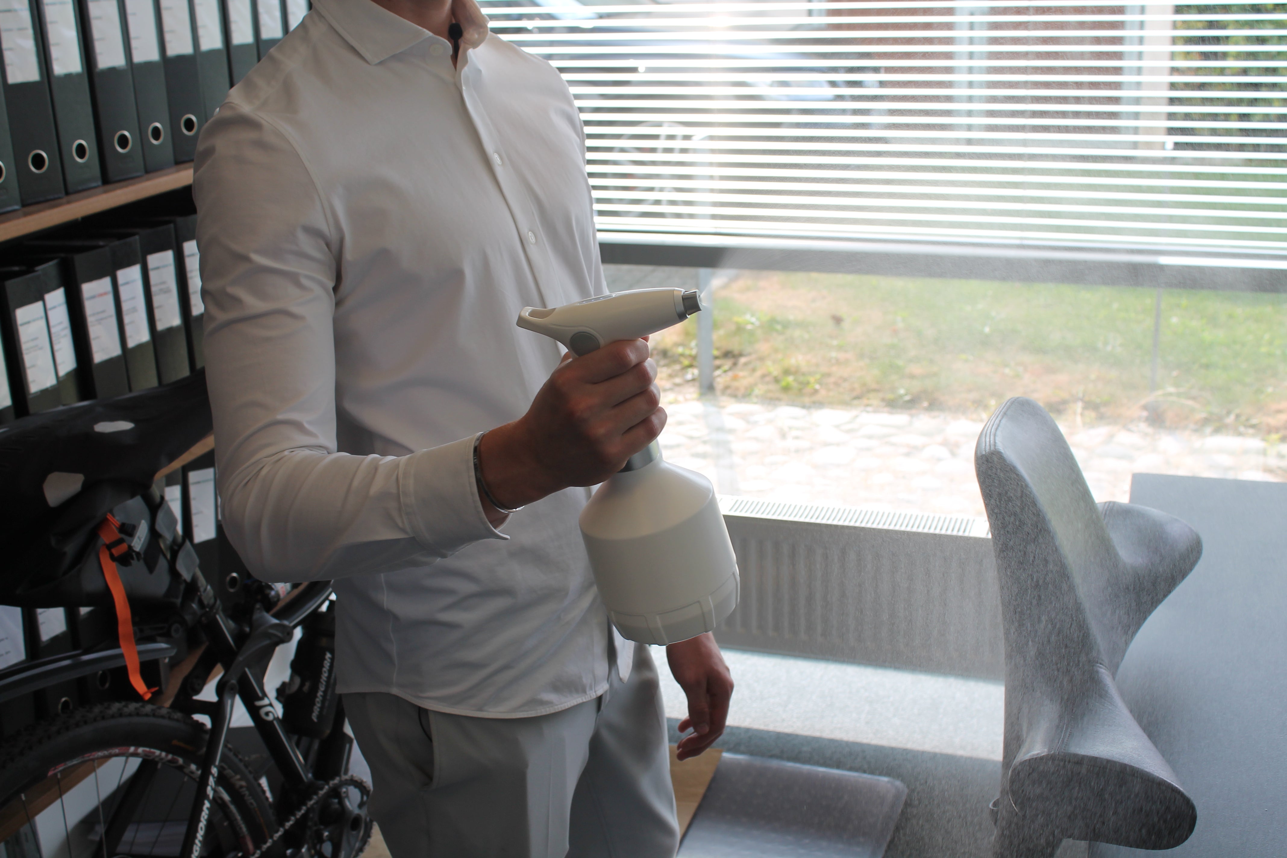 På billedet ser man safesprayer adjust elektrisk forstøver anvendes til at forstøve et desinfektionsmiddel ud på skrivebord. Sprayeren bliver anvendt af en mand med hvid skjorte.