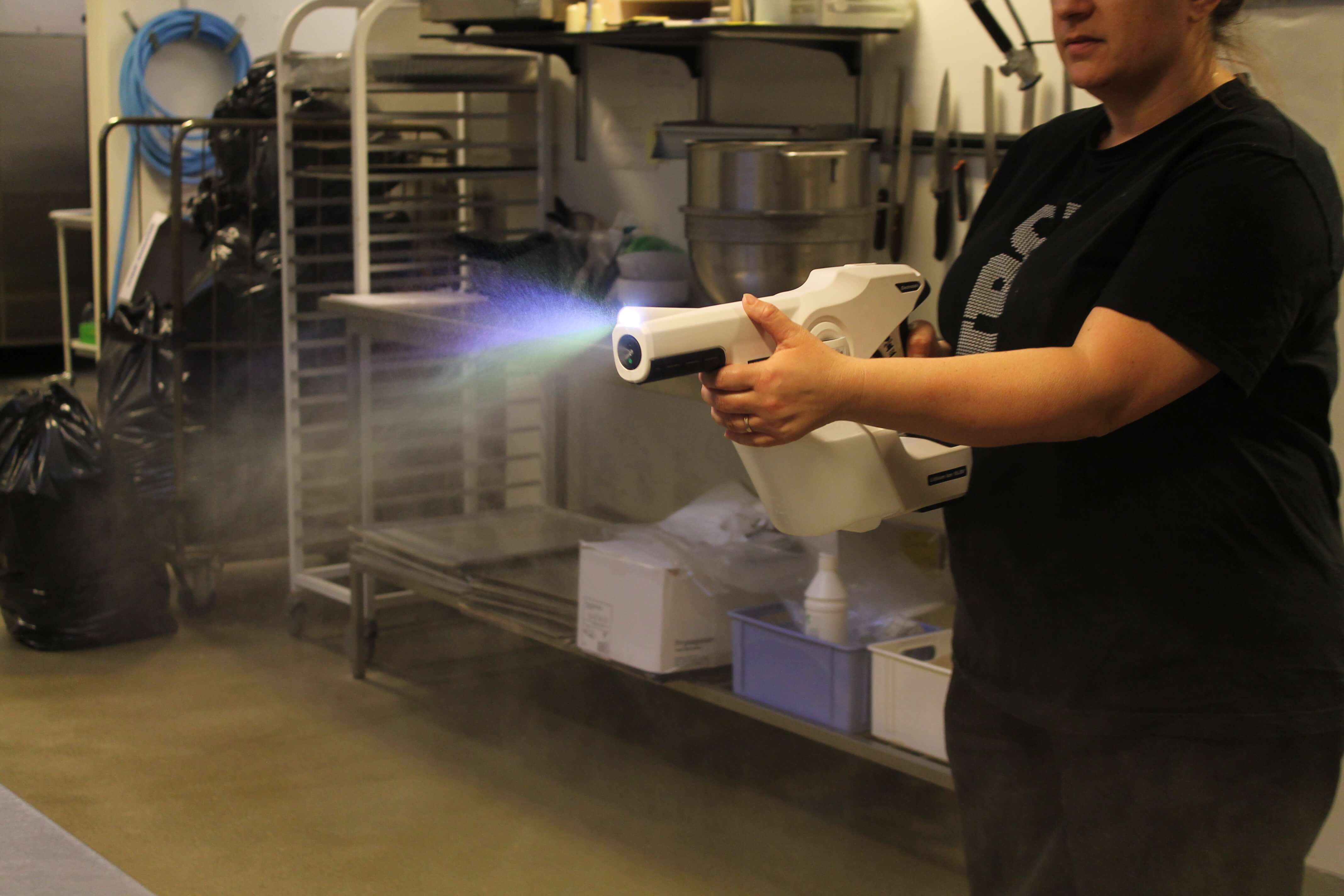 På billedet ses i forgrunden en Protexus sprayer som er en elektrostatisk forstøver til desinfektion. Den holdes af en kvinde med en sort trøje og der kommer væske ud af dysen. I baggrunden ses et køkkenmiljø.